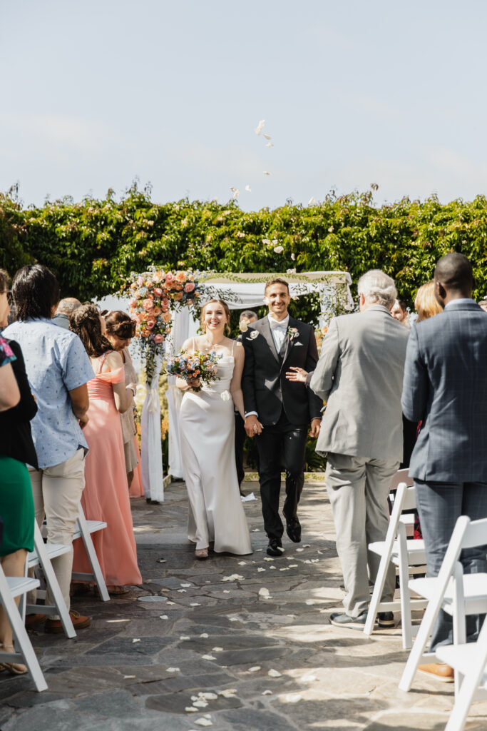 Bride and groom walk down the aisle smiling after their outdoor wedding ceremony under a floral arch.