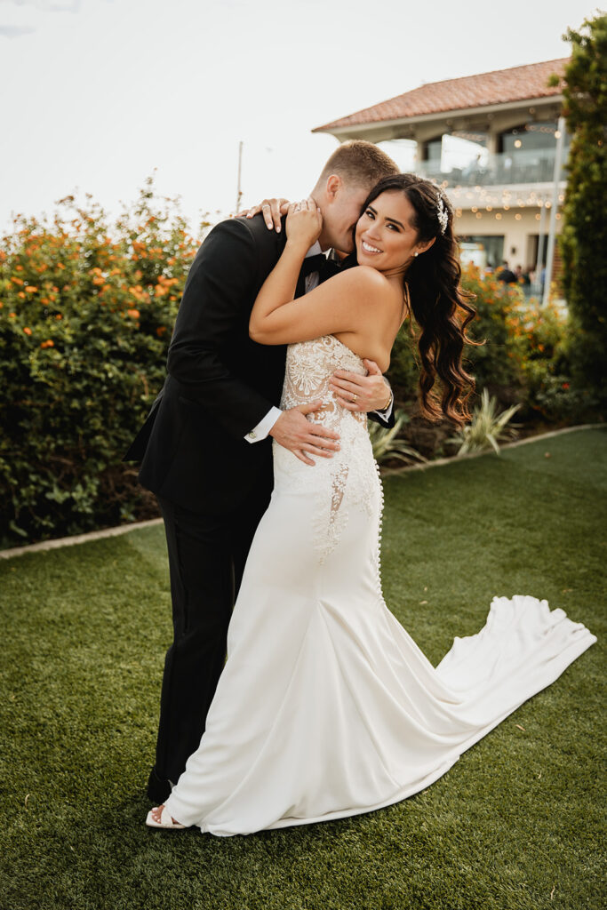 Bride and groom share an intimate embrace on a lush lawn with a Spanish-style building in the background.