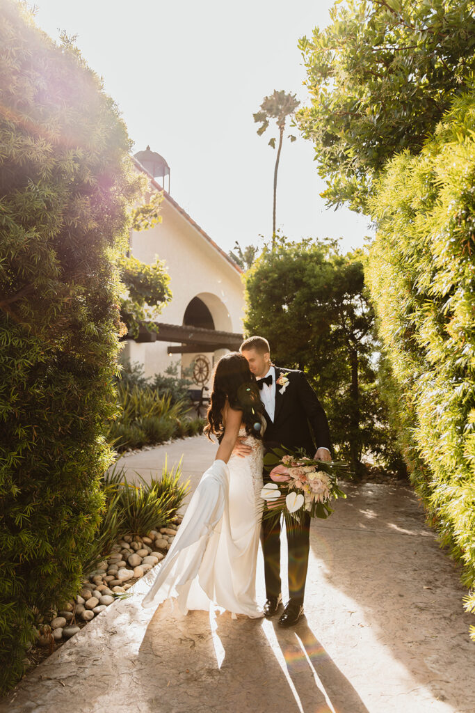 Bride and groom share a kiss under the golden glow of sunset, surrounded by greenery and soft light.