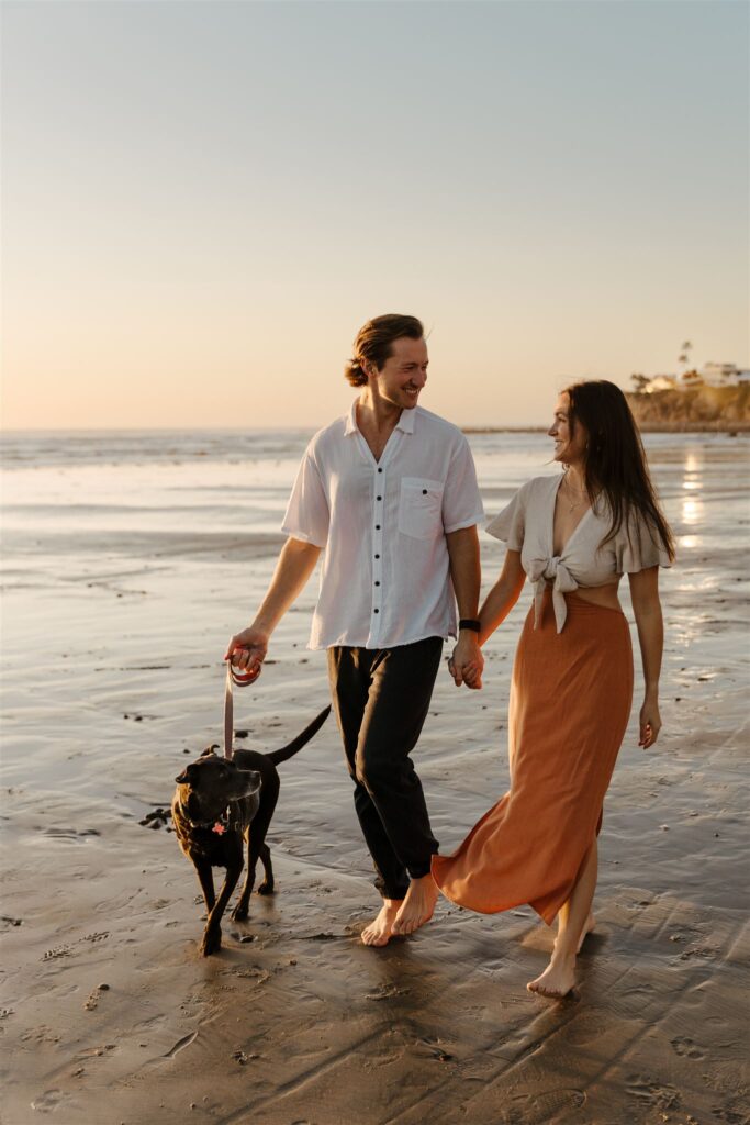 Happy couple walking their dog along a beach during sunset in San Diego.