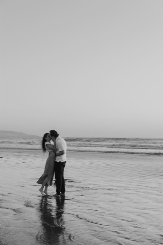 Black-and-white photo of a couple kissing on the beach with their reflections in the wet sand.