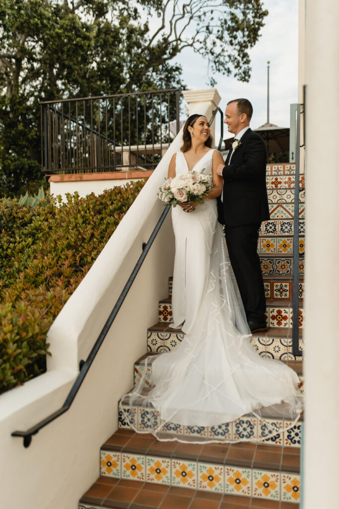 Bride and groom pose together on colorful tiled stairs, holding a bouquet of soft pink and white roses.