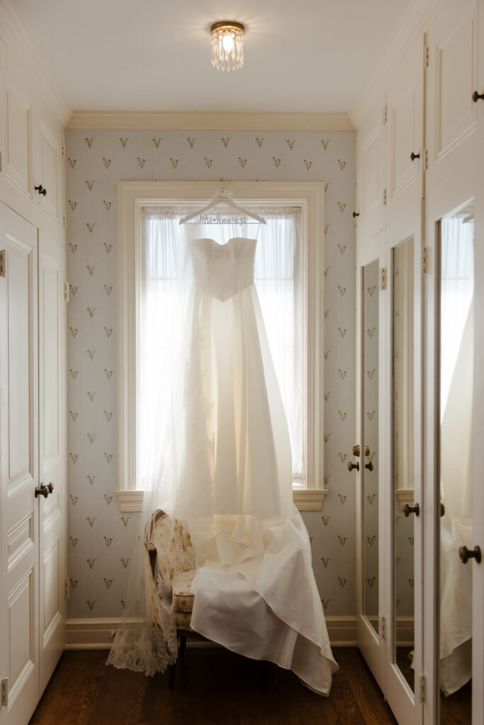 Elegant strapless white wedding gown displayed in a classic bridal suite with natural light streaming through the window.