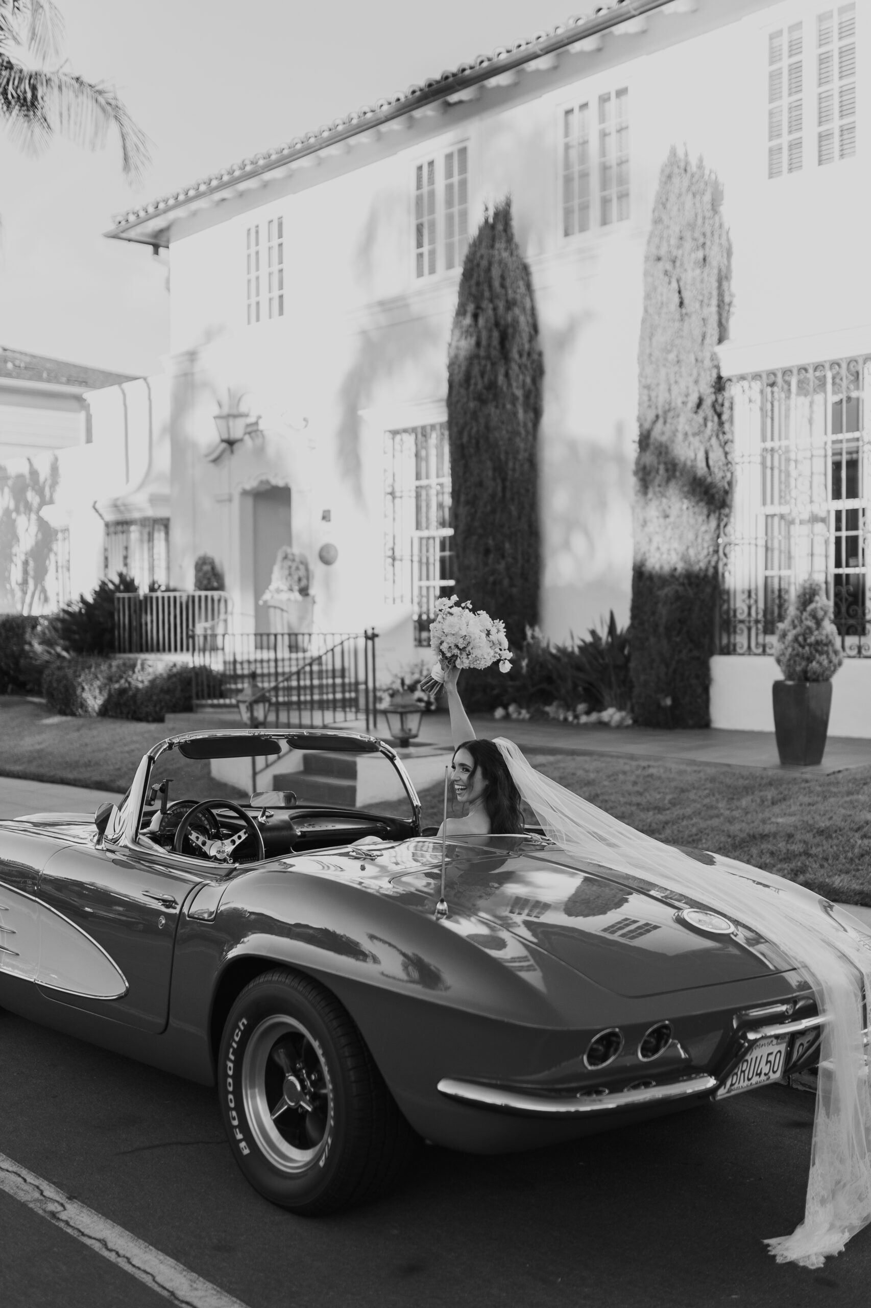 Bride in a vintage convertible holding her bouquet with a veil flowing gracefully down the back of the car.