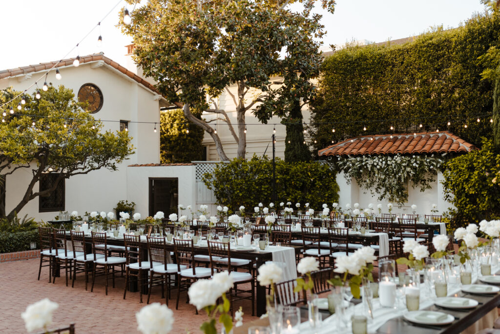 Elegant outdoor reception setup with long tables, white floral arrangements, and bistro lighting under a canopy of trees.