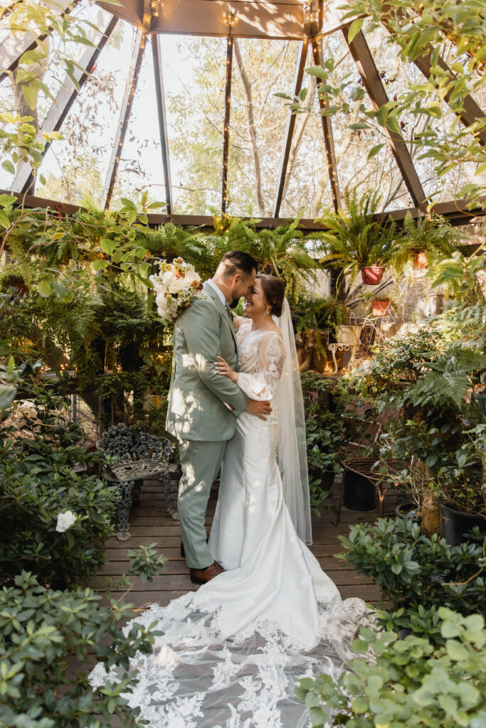 Bride and groom share a romantic kiss inside a lush greenhouse filled with greenery and soft natural light.