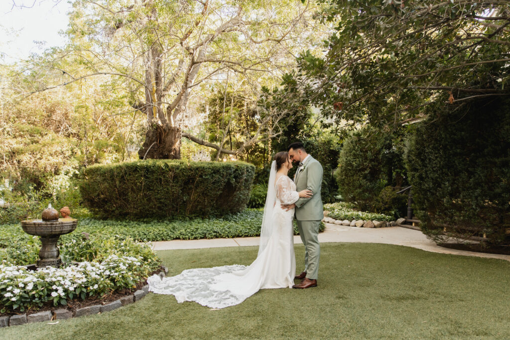 Bride and groom stand forehead-to-forehead in a beautifully manicured garden surrounded by trees and flowers.