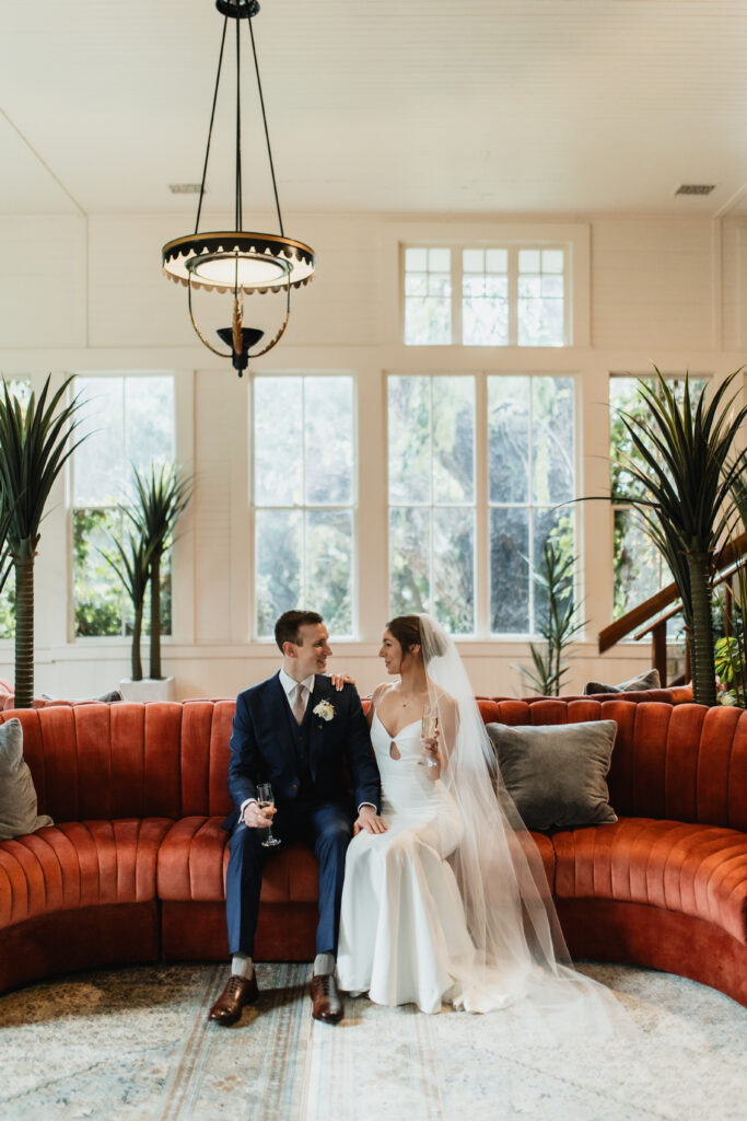 Bride and groom share a quiet moment seated on a vibrant red velvet sofa in a light-filled reception venue.