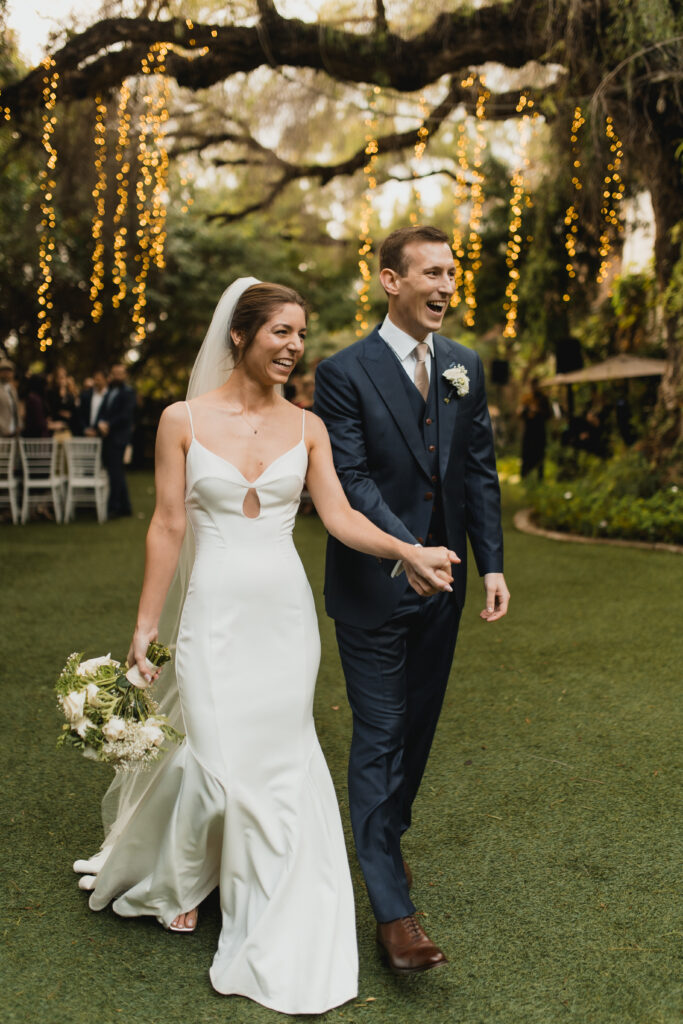 Bride and groom joyfully walk hand-in-hand down the aisle surrounded by glowing string lights in a garden setting.