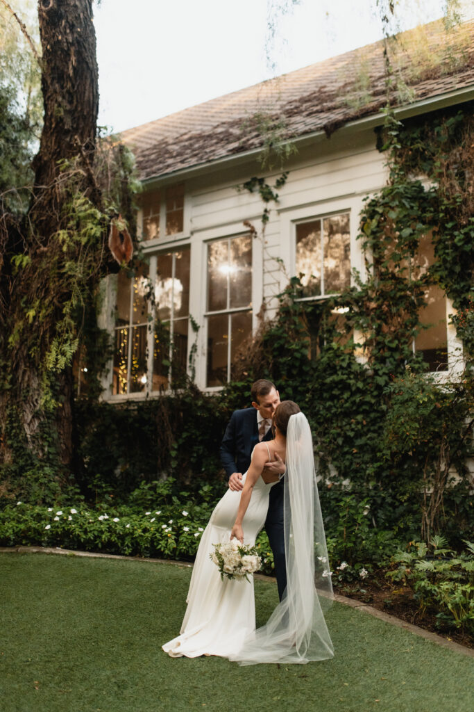 Bride and groom share a romantic kiss in a lush garden with a historic building covered in ivy as the backdrop.