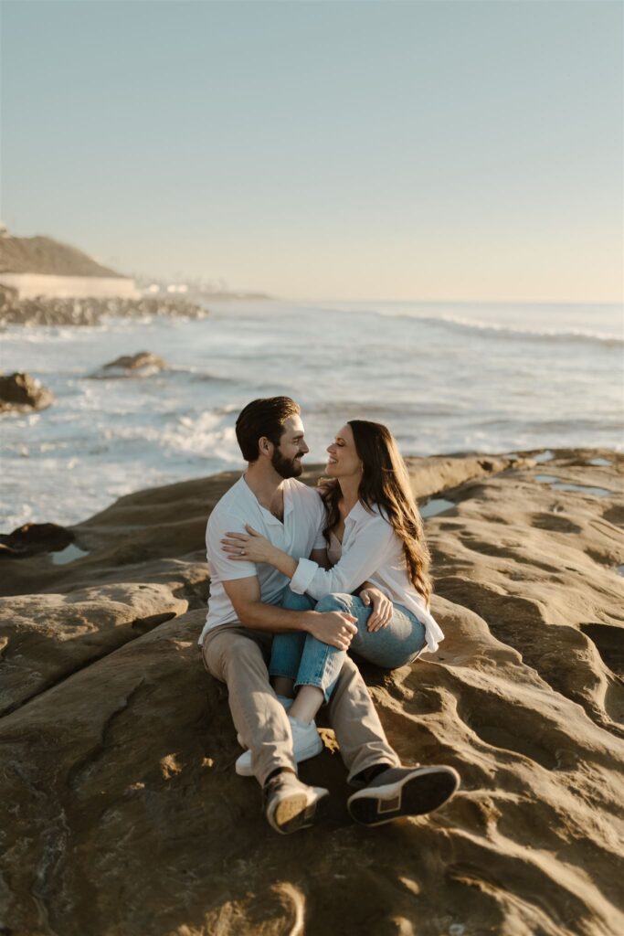 Couple sitting on rocks near the ocean, gazing lovingly into each other's eyes.