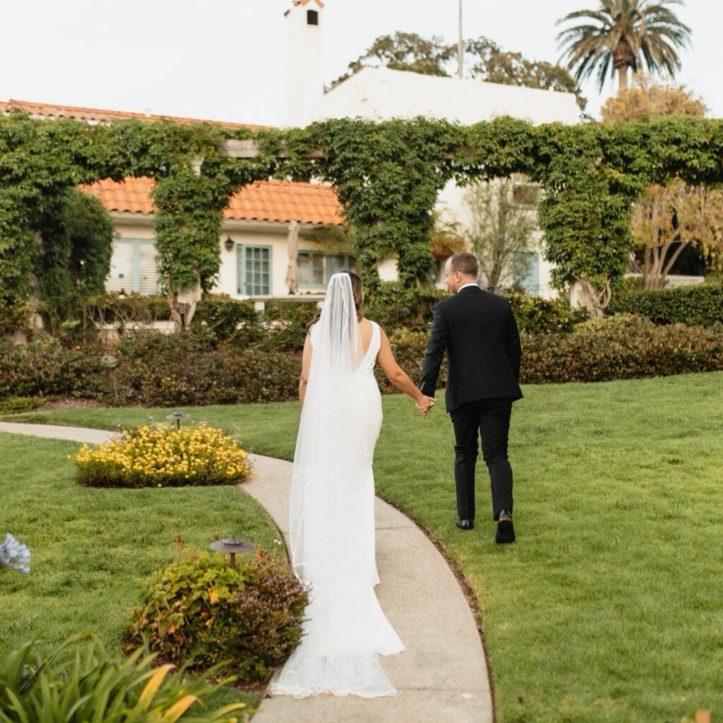 Bride and groom walk hand-in-hand through a lush garden with a romantic Spanish-style backdrop.